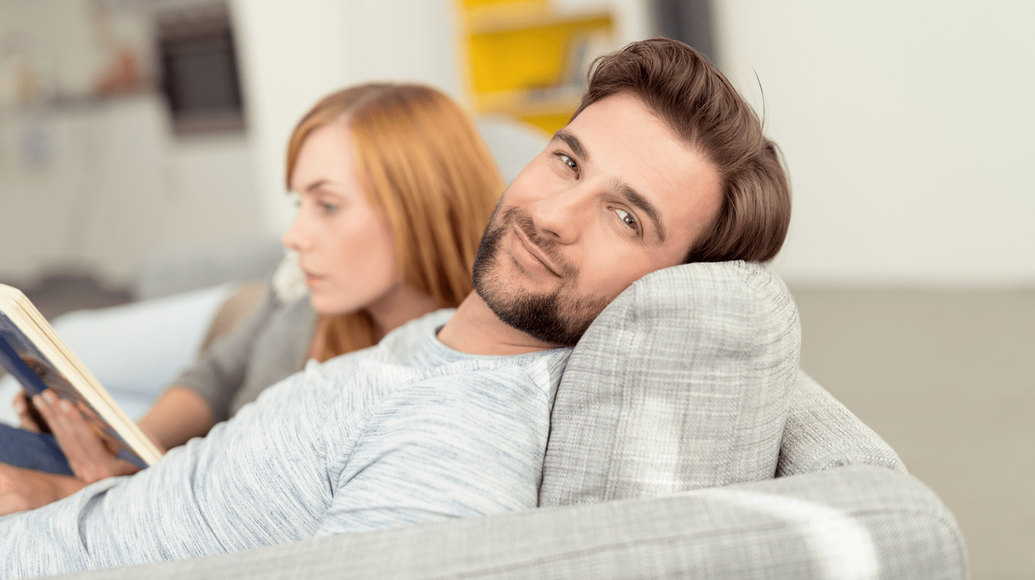 Pärchen. Ein Pärchen sitzt auf dem Sofa, der Mann schaut in die Kamera und die Frau liest ein Buch.
